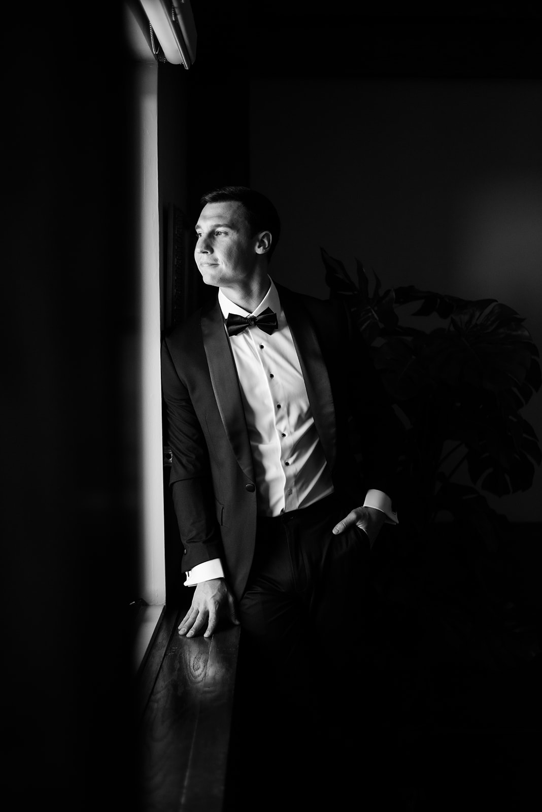 Black and white film portrait of the groom looking thoughtfully out a window before the wedding.